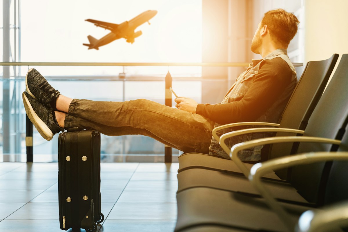 A traveler waiting with luggage inside an airport terminal as a plane takes off